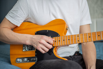 A man in a white T-shirt plays a natural-colored electric guitar sitting on a bed in close-up,...