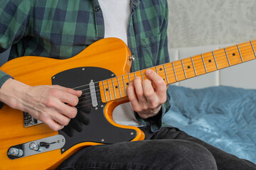 A man in a plaid shirt plays a natural-colored electric guitar sitting on a bed in close-up,...