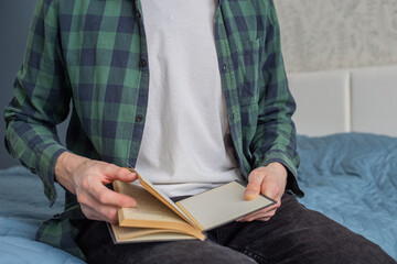 A man in a plaid shirt is reading a book, sitting on the bed, close-up, in selective focus. A man reading a book in a cozy atmosphere. A man is leafing through an old book with yellow pages.