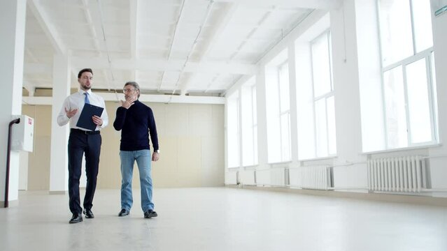 Realtor talks to client walking through an empty rental space, front view. Realtor and businessman walk around an office space for rent and talk
