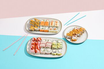 Big sushi set on ceramic dishware with coloured chopsticks. Trendy set of maki sushi in minimal style. Modern japanese menu concept. Maki sushi assorted on coloured background.