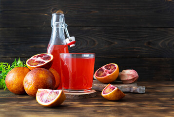 A transparent glass of Sicilian orange juice on a wooden stand. A bottle of juice and red oranges in the background. Wooden background, space for text