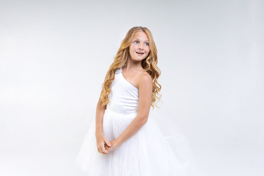 Beautiful cute girl in white dress with long wavy hair posing in studio on background. Caucasian school age girl model