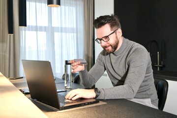 Portrait of happy positive guy, young cheerful man is enjoying internet online shopping, purchasing on his laptop computer and holding credit bank card in hand, sitting at home, smiling, entering