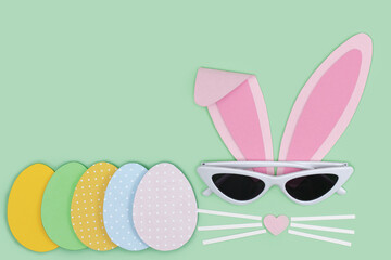 Easter greeting card. Bunny rabbit face made of pink paper with sunglasses and painted Easter eggs cutting out paper on blue background. Happy Easter minimal concept with copy space. Paper Crafts