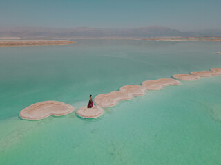 Aerial shoot of girl in long romantic red dress walking on Salty islands of Dead Sea among mirror smooth water with Jordan mountains on the horizon. Seascape, Nature background. Israel