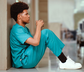 Emotional stressed intern doctor sitting against wall on floor in hospital corridor. Distraught...