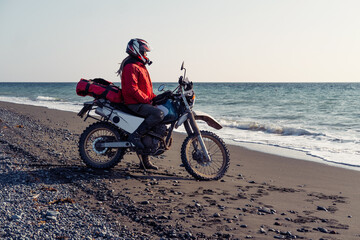 Woman riding enduro motorcycle standing on the shore near waves of the ocean. Female solo...