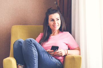  Beautiful  Smiling  Woman   sitting at  cozy chair and  looking at the Phone  reading a Message ,checking social media and News, ordering delivery  connected to high speed internet