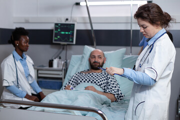 Middle aged man recovering lying in hospital bed attended by team of doctors after surgical intervention. Doctor with stethoscope reading medical chart while nurse is monitoring patient vitals.