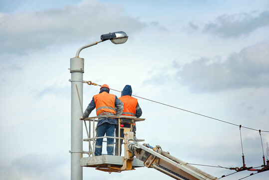Workers in aerial platform paint street lighting pole. Men in cradle painting lamppost with paint brush. Cherry picker with workers at height, risky job.  Paint and renovate street lightning pole