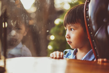 Kid looking out window. Adorable little caucasian kid boy stand still near window glass and looking to outside alone during bright sunny day. Little child reflecting bored and loneliness at home