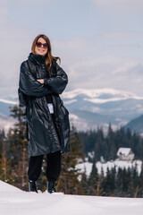 Fototapeta na wymiar A young girl in sunglasses and a black jacket at a ski resort against the backdrop of mountains