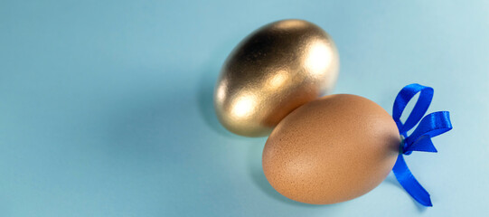 banner with Brown egg with blue ribbon and golden egg. Happy Easter concept. Two eggs with ribbon on a pastel blue background with copy space.