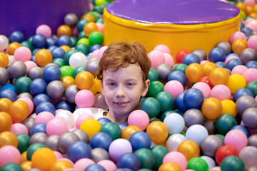 Fototapeta na wymiar A teenage schoolboy with a pale face and red hair sits in a ball pool and smiles slyly.
