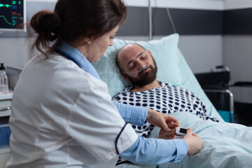 Doctor putting pulse oximeter on finger of sleeping patient with nasal cannula connected to oxygen...