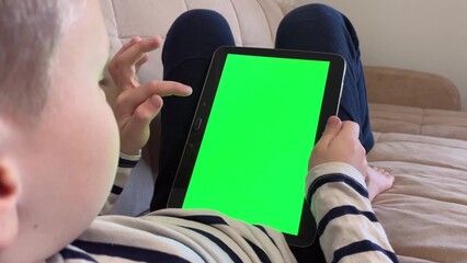 Child lying on the couch and using a digital tablet PC with green screen, back view. Boy holding a tablet with green screen in hand