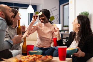 African american woman playing guess who game with coworkers, celebrating with drinks after work. Office colleagues enjoying charades pantomime play with sticky notes at party.
