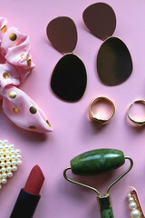 Pink scrunchie, gold earrings and rings, blush, various hair barrettes, lipstick and jade roller on bright pink background. Flat lay.