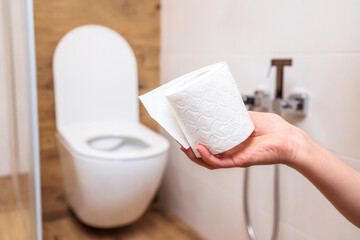 A woman's hand holds a roll of white toilet paper close-up. Bathroom background