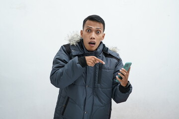 Asian man wearing winter jacket over isolated white background. Shouting and looking at the camera while pointing at his smartphone received bad news