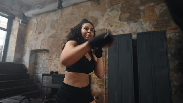 Woman with muscular body kicks punching bag with fists