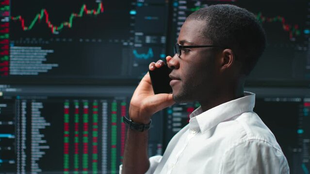 African-American trader talk on smartphone standing over multiple monitors with charts and data