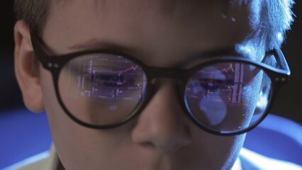 Close-up of the face of a boy with glasses sitting at a computer and looking at an online stock market chart showing bitcoin currencies. The glasses reflect the bitcoin graph. In real time