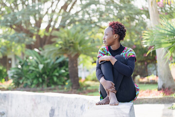 Young relaxed African girl sitting cross-legged on a low wall in the park waiting for her friends to come over and do something together