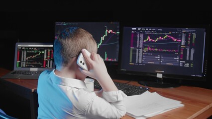 A rear view of a boy sitting at a computer and looking at an online stock market chart showing bitcoin currencies. the boy is talking on the phone. In real time. Cryptocurrency. Investors