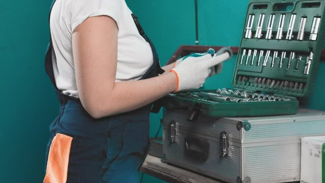 A female mechanic in overalls and gloves choice a nozzle for a ratchet wrench in a box with tools and repair of cars engine. Hands close-up.