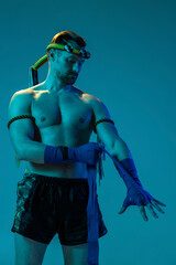 Half-length portrait of muscled man, thai boxer in sports uniform posing isolated on blue studio background in neon. Sport, muay thai, competition, fight club concept