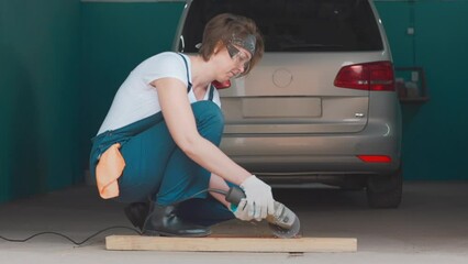 A young caucasian woman in coveralls, gloves and goggles is working with an angle grinder. Slow motion. Car repair shop. - Powered by Adobe