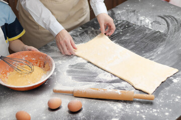 The female hands of the cook roll out puff pastry for baking on a black kitchen table with a wooden rolling pin. Chicken eggs, flour and a bowl of dough. selective focus