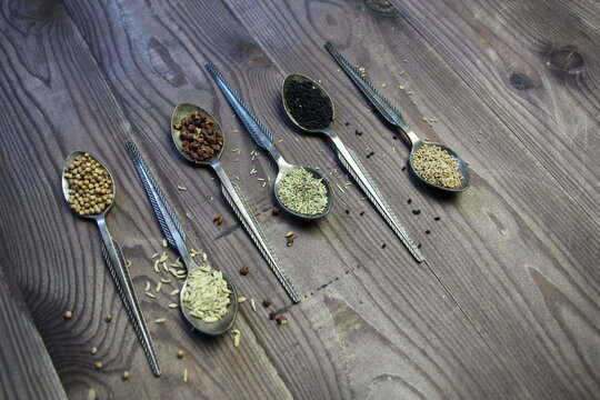 Metal vintage spoons with spice seeds of anis, nigella, ajowan, coriander, rosemary, Sichuan pepper on brown wooden table