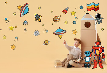 Cute little boy with books, toy and cardboard rocket pointing at drawn stars and planets on color...