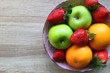 Pink bowl filled with fresh apples, oranges and strawberries on wooden table. Flat lay.