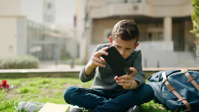 Blond child playing video game by smartphone sitting on grass at park