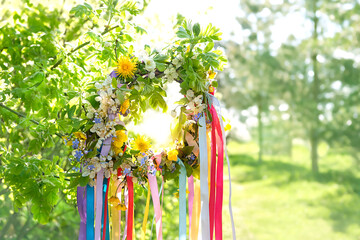 Spring flower wreath with colorful ribbons in garden, green natural background. floral decor, Symbol of Beltane, Wiccan Celtic Holiday beginning of summer season. pagan witch traditions, rituals. - obrazy, fototapety, plakaty