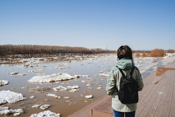 The girl stands with her back watching the flood on the river, the ice floats in pieces on the...
