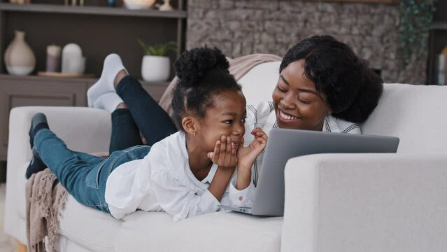 Happy parent african mother and cute child daughter looking at laptop screen talking having fun video call choosing in internet store using computer app game at home relaxing laughing lying on sofa