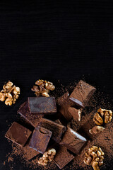 A pile of dark broken chocolate sprinkled with cocoa powder and walnuts on a dark background. Copy space
