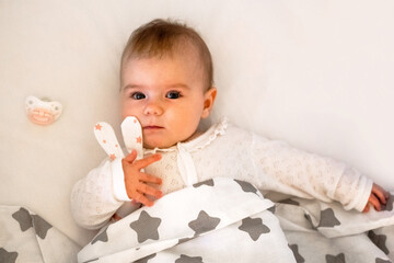 Adorable Caucasian baby girl six months old lying on white bed with toy of hare in her hand.
