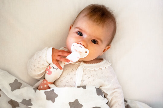 Candid portrait of cute Caucasian baby girl lying on white bed with pacifier and toy of hare.