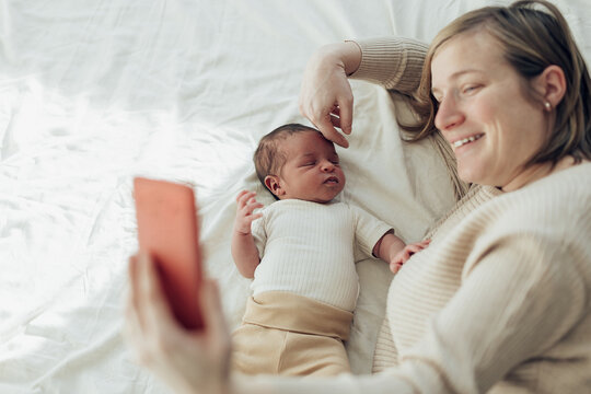 happy mum lying in bed with newborn baby making a video mobile phone call with family and friends