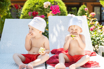 happy boys twins in panamas and swimming shorts eating apples on sunbeds with sunscreen on face
