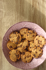 Pink plate filled with chocolate chip cookies on wooden table. Flat lay.