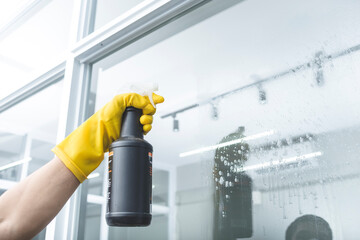 An anonymous man wearing yellow gloves sprays the surface of a window with glass cleaner.