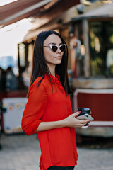 Elegant attractive european woman walking outdoors with coffee. Woman in sunglasses wearing red blouse while listening to music.