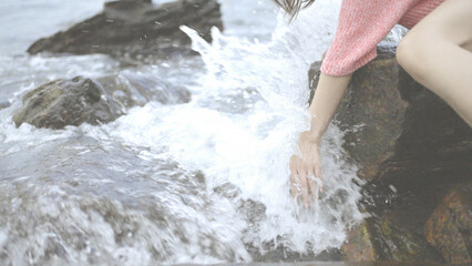Girl sitting on the rocks. Young woman's hand touching the water in the sea, catching the wave with her hand. Splashing Waves at Blowing Rocks.